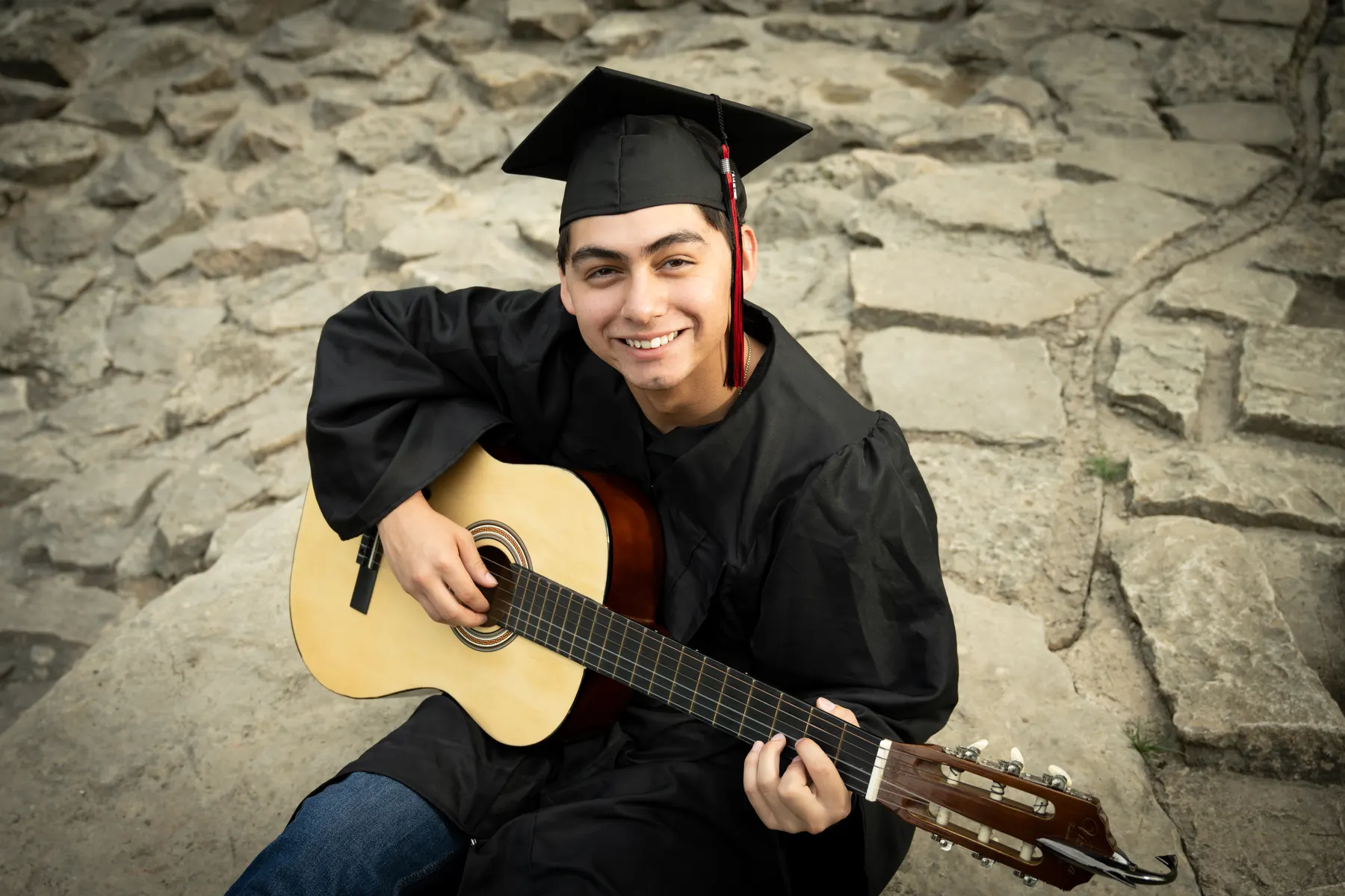 A smiling graduate in a black cap and gown holds an acoustic guitar on stone steps