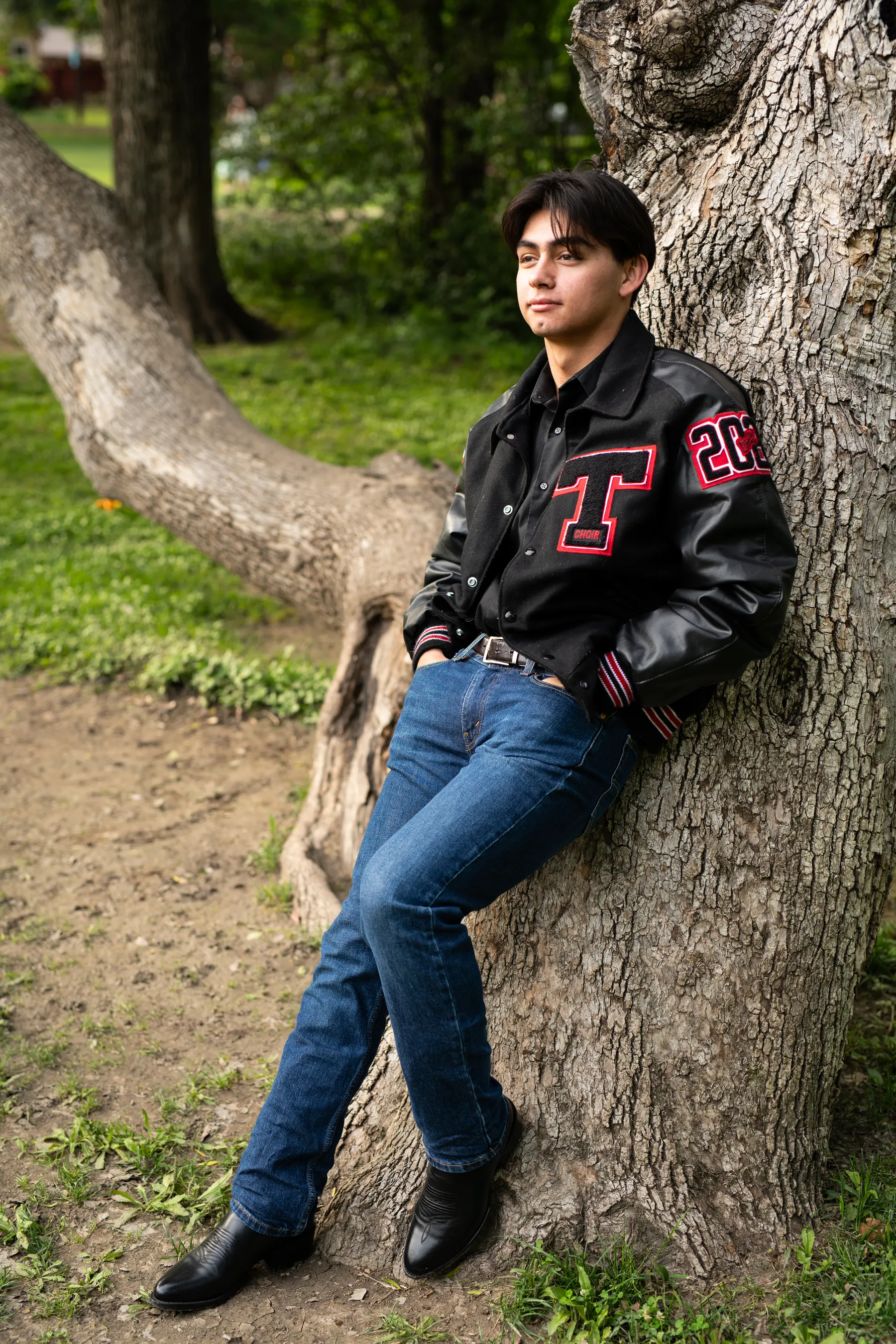 A senior in a black and red letterman jacket and jeans leans against a large tree