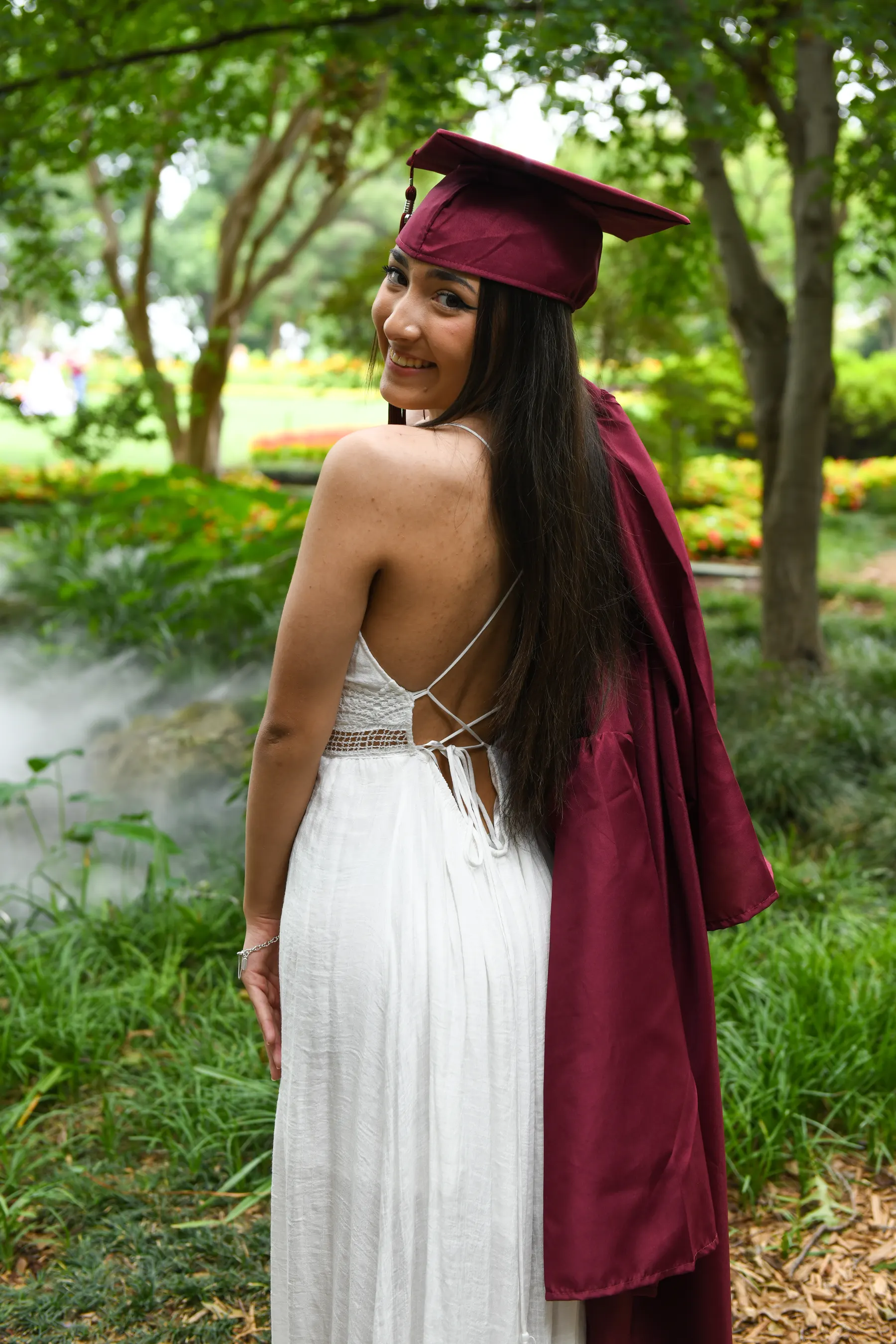 A graduate in a maroon cap looks over her shoulder, her gown draped over a white dress in a leafy garden