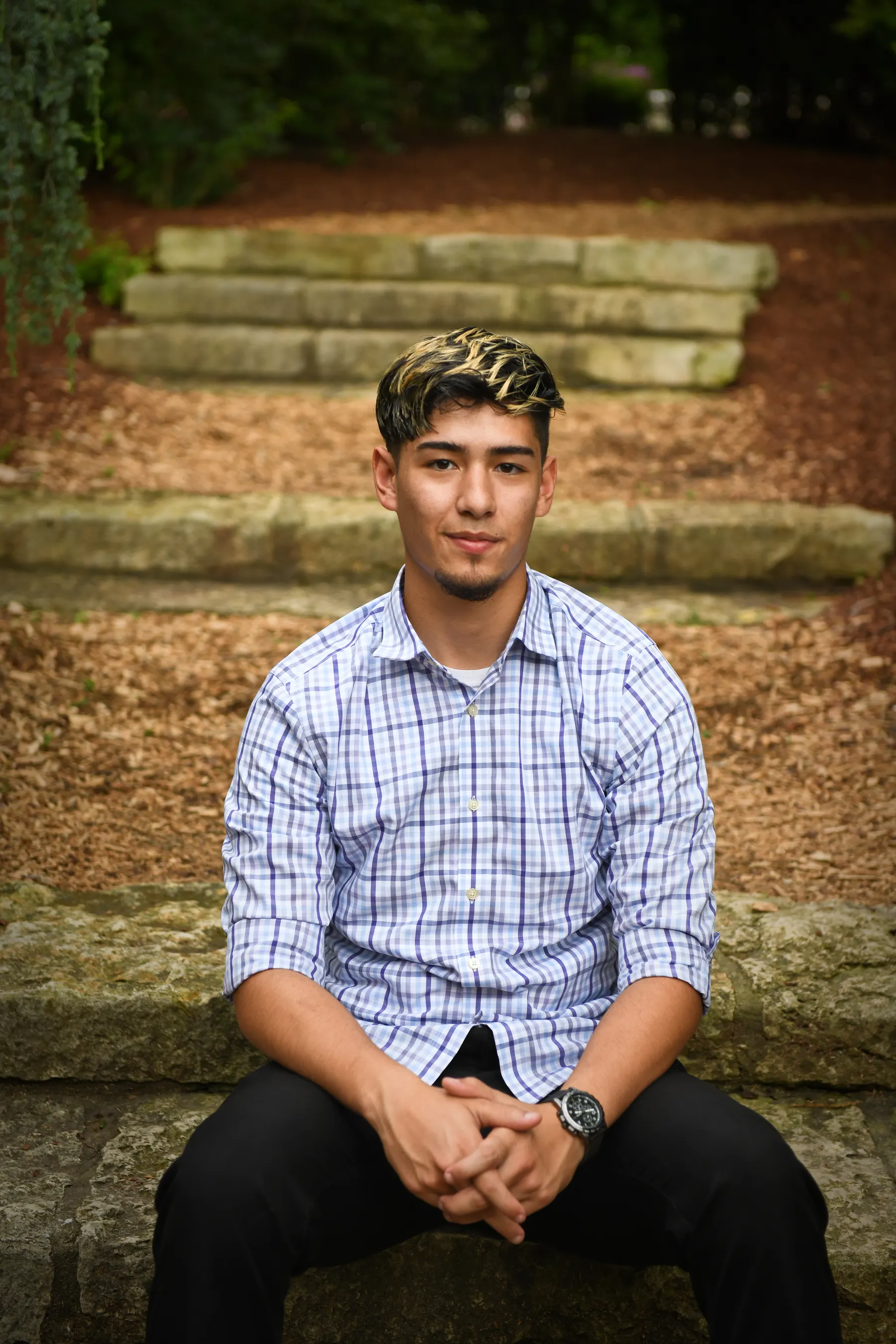 A senior in a blue checked shirt sits with his hands folded on stone garden steps
