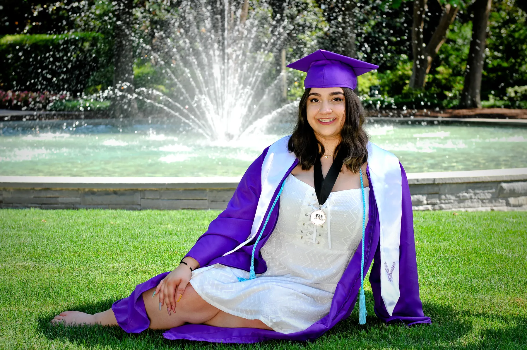 A graduate in a purple cap and gown sits on the lawn in front of a garden fountain
