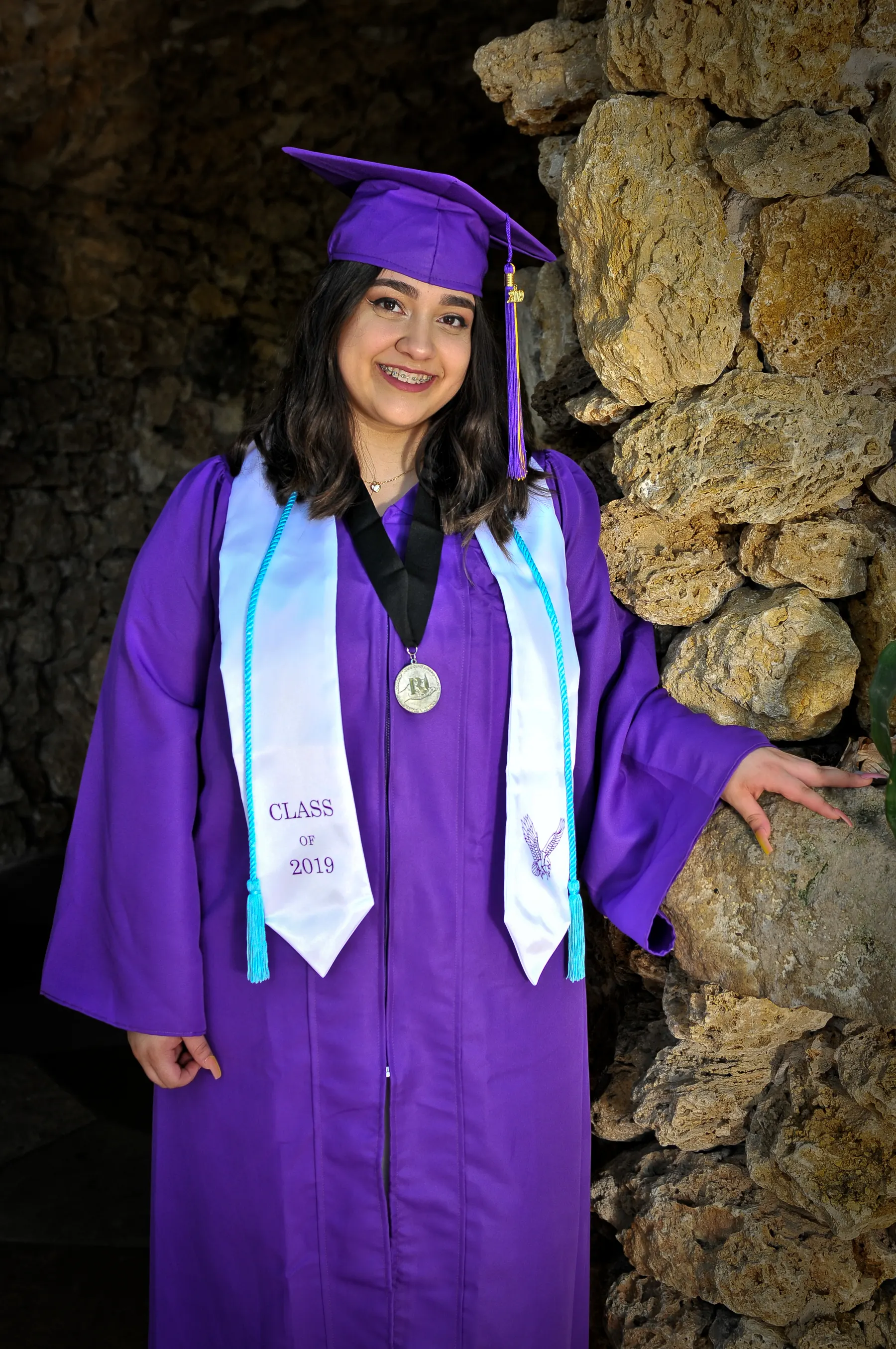 A graduate in a purple cap and gown with a white stole smiles beside a stone archway