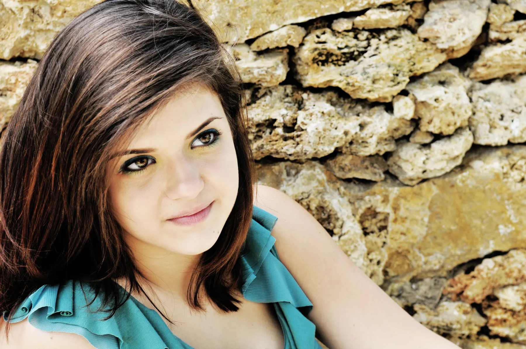 A senior in a teal top looks up at the camera beside a textured stone wall