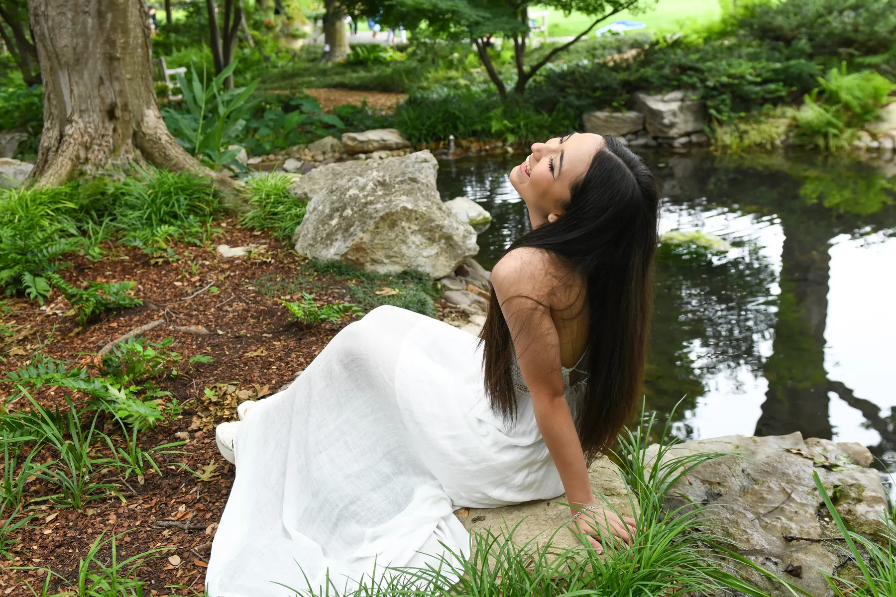 A senior in a flowing white dress laughs while seated on a rock beside a garden pond